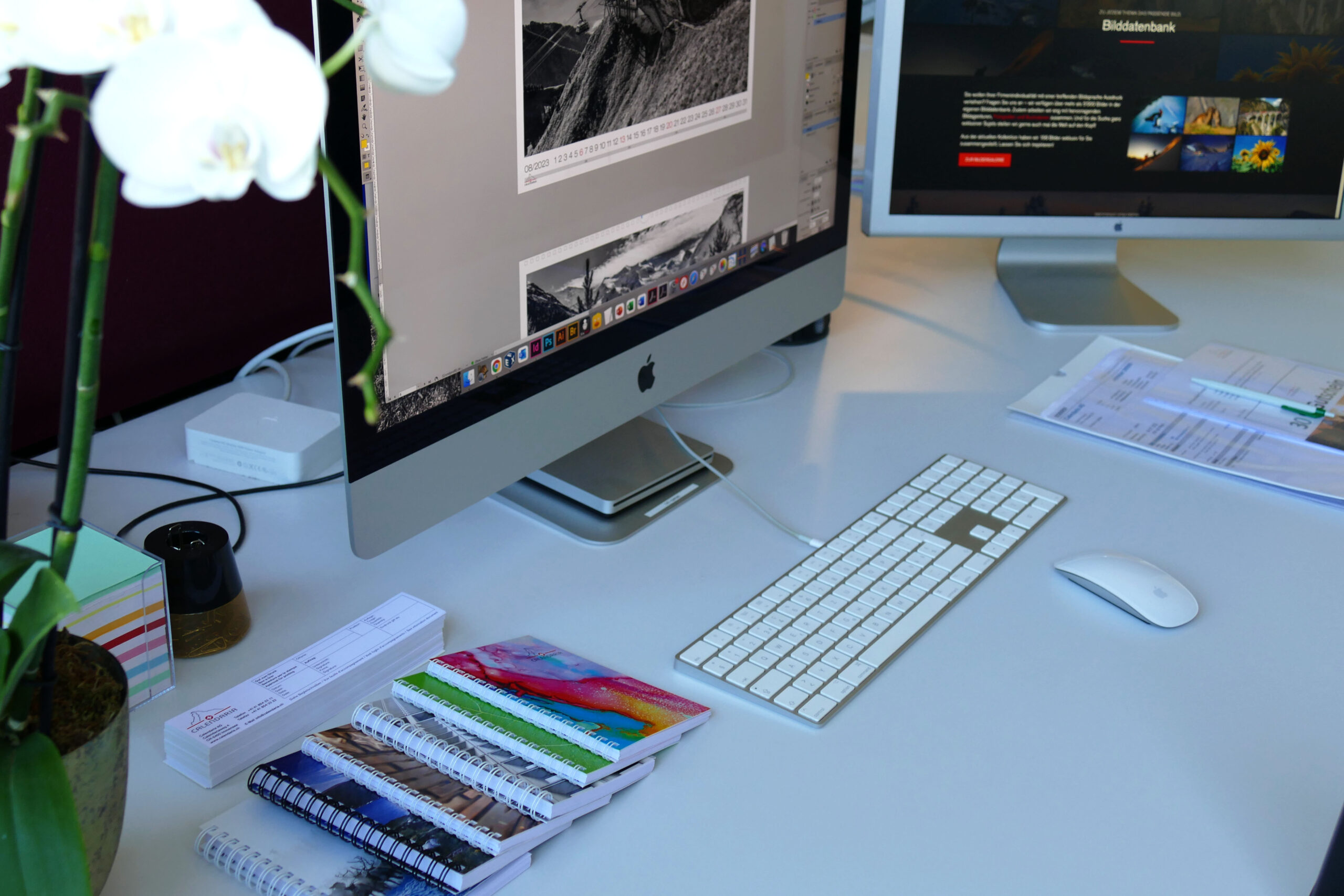 Grafik Arbeitsplatz mit zwei Apple Bildschrimen, Tastatur, Maus, Blumen und Wiro gebundene Druckprodukte